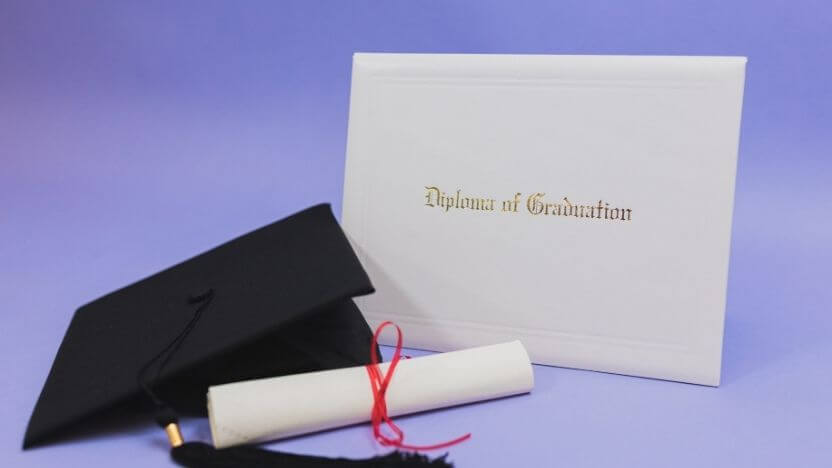 diploma of graduation folder with shiny gold letting sitting next to black graduation cap and rolled up diploma with red ribbon